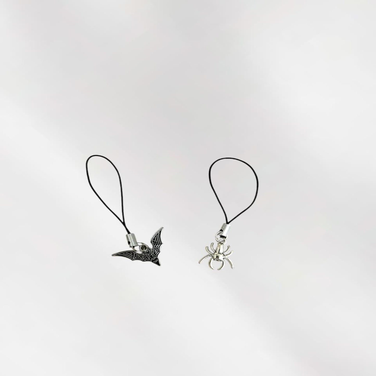 Two phone charms, one with a bat design and the other with a spider design, on black strings against a white background.