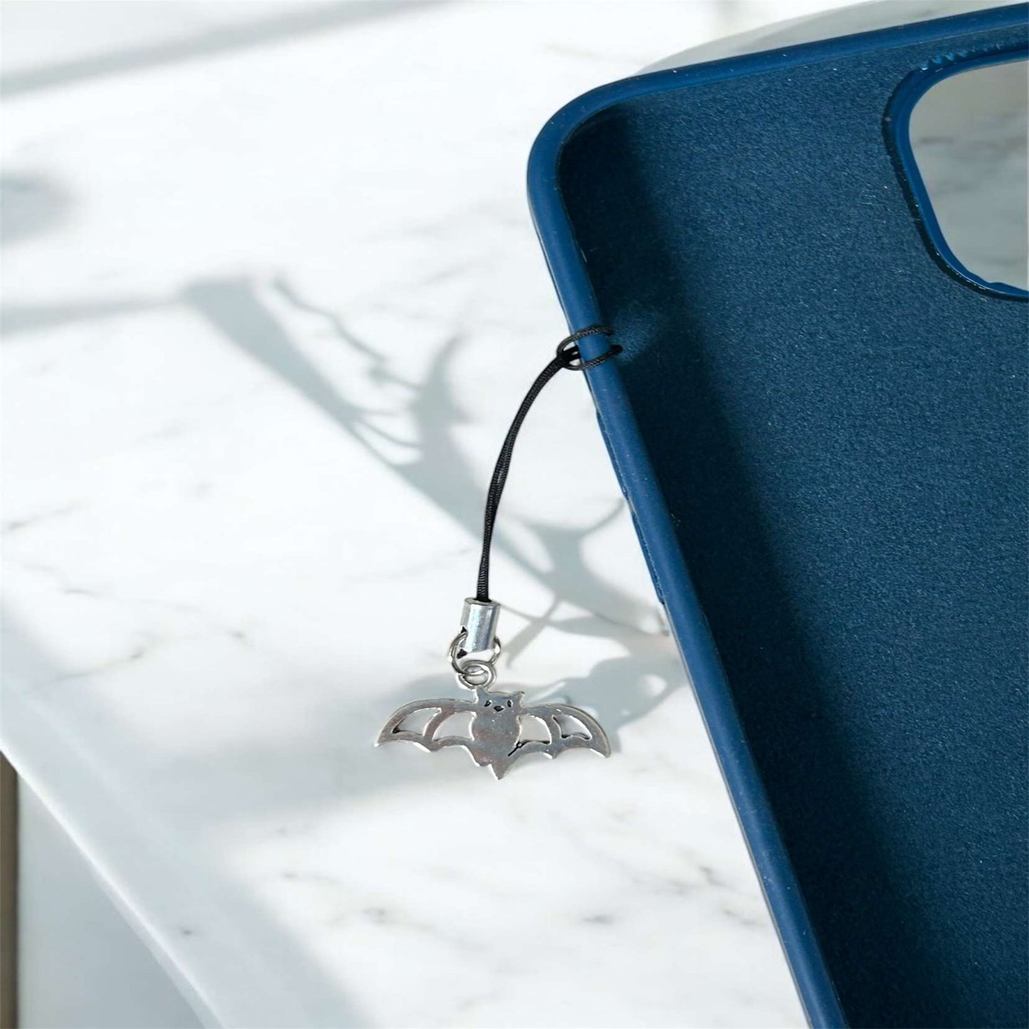 Silver bat-shaped phone charm attached to a blue phone case on a white surface. - Thecharmingcharms09