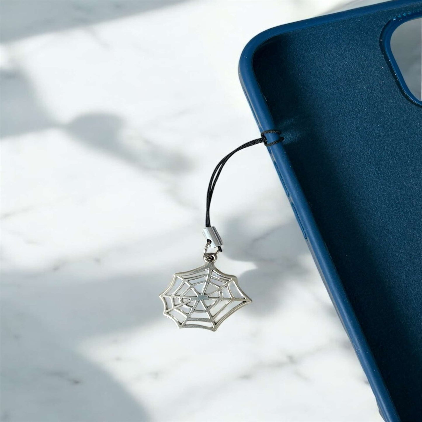 Silver spider web charm on a black cord attached to a blue phone case on a light marble background - Thecharmingcharms09